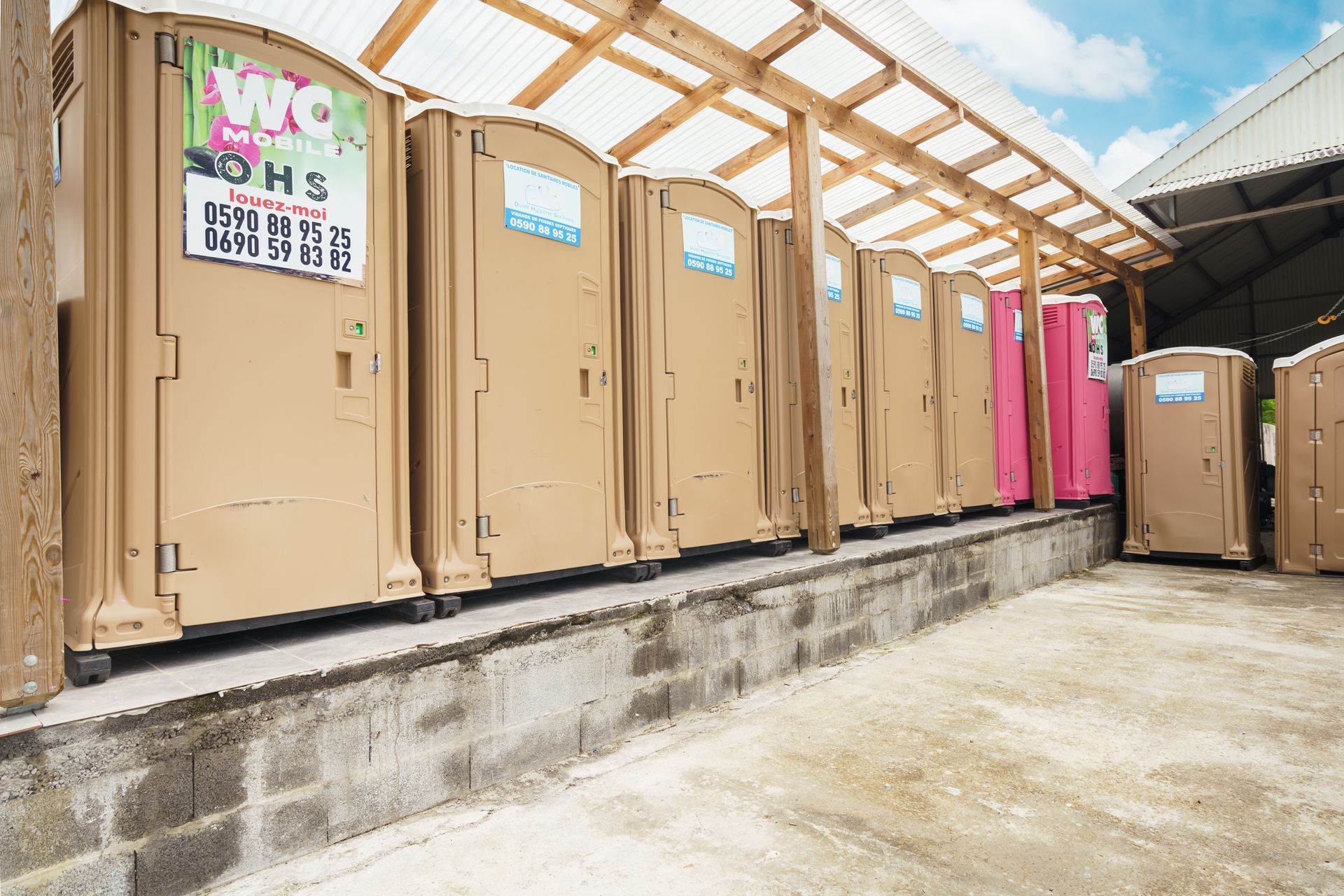 Une rangée de toilettes portables beiges et roses borde une allée en béton sous un auvent en bois.