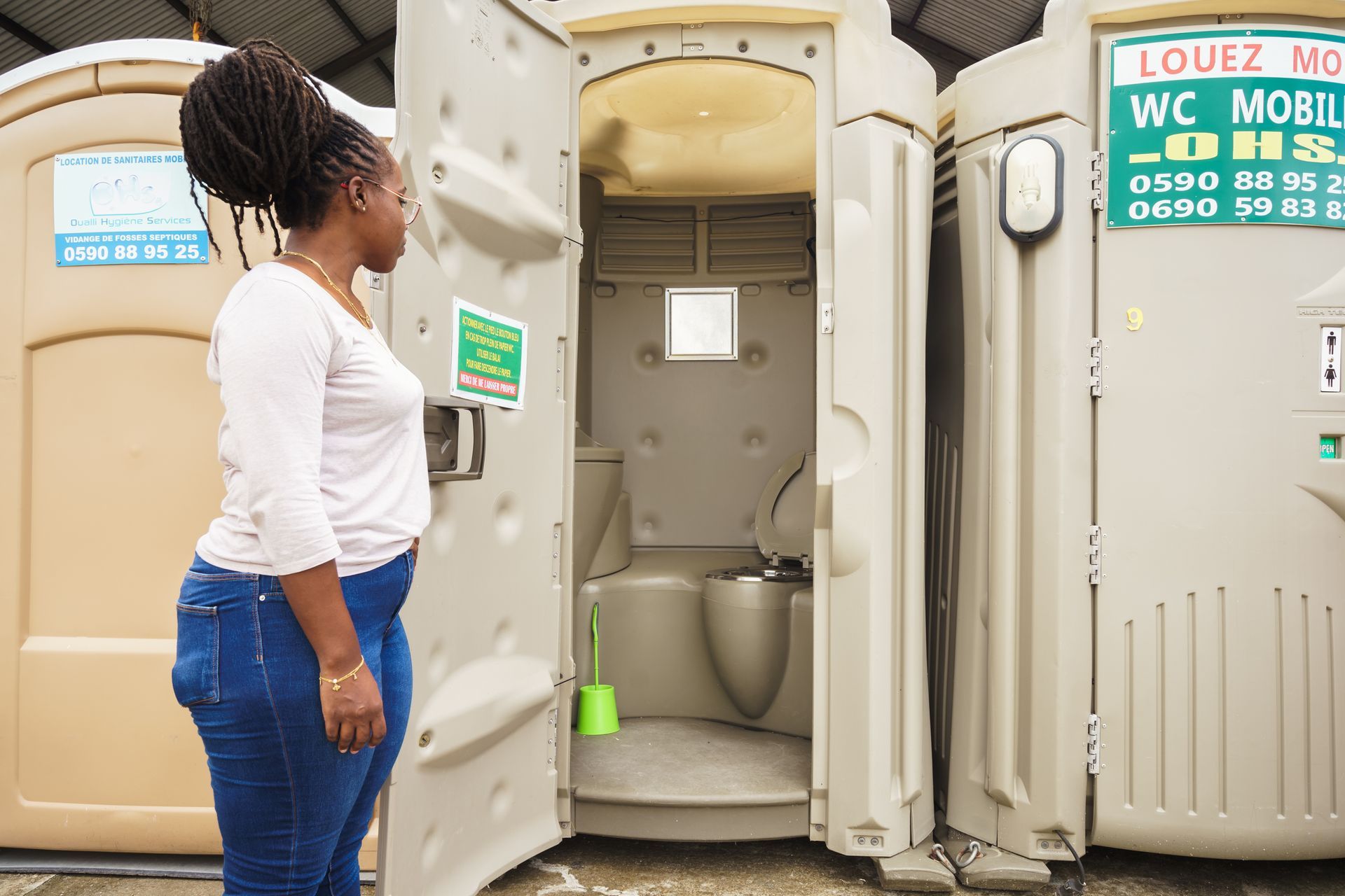 Une femme se tient devant des toilettes portables, la porte ouverte.