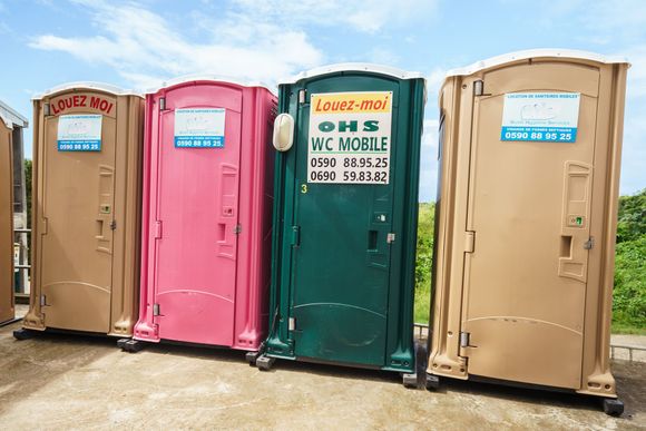 Quatre toilettes portables colorées alignées à l'extérieur, chacune avec une pancarte sur la porte.