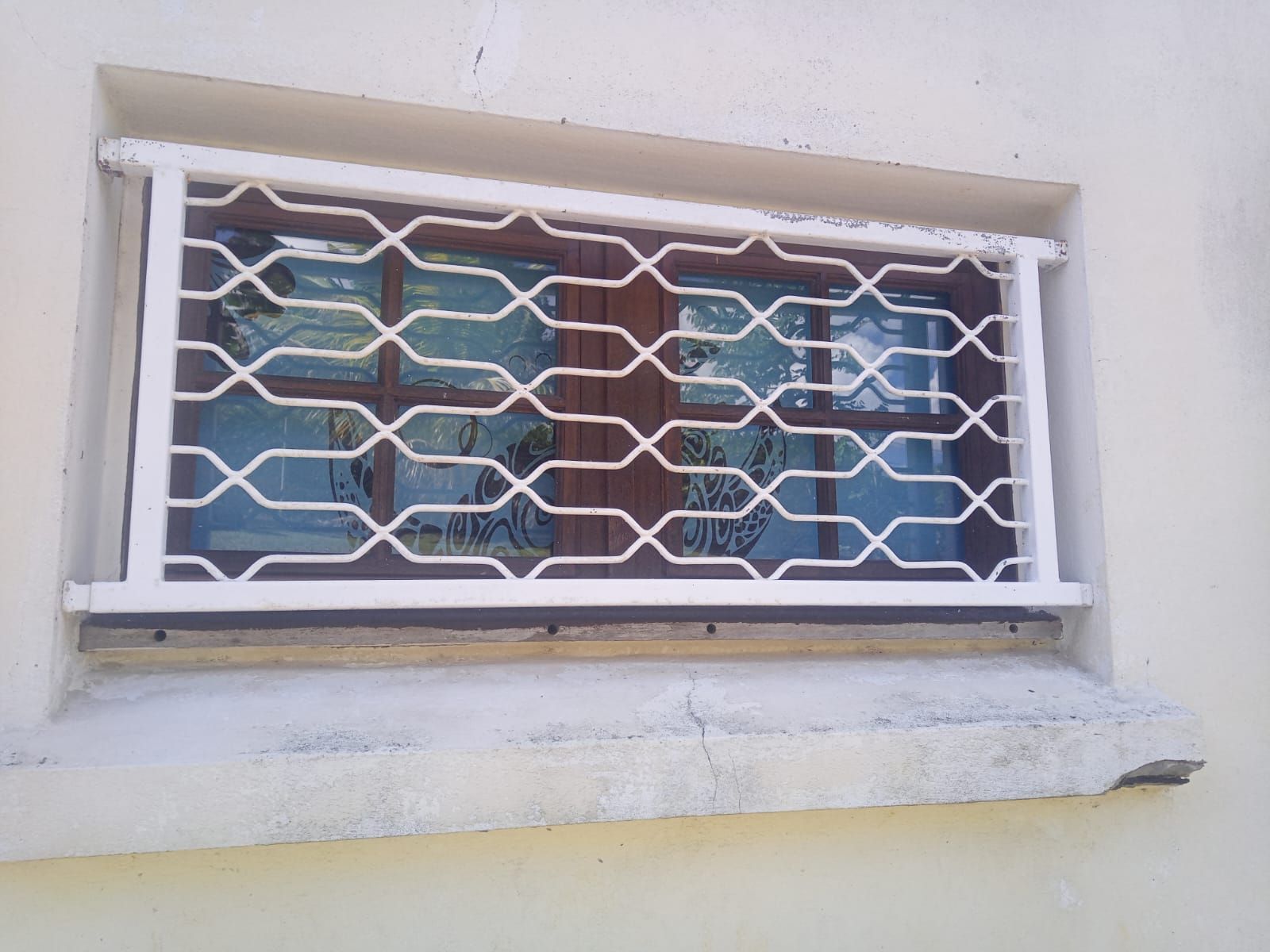 Une petite fenêtre rectangulaire à cadre marron, recouverte d'une grille de sécurité en métal blanc.