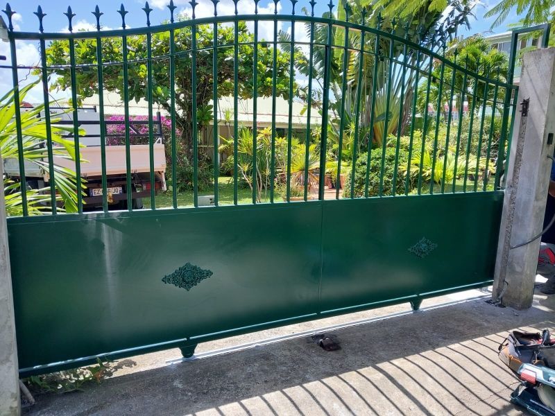 Un portail d'allée en métal vert avec des barreaux verticaux et des panneaux centraux décoratifs.