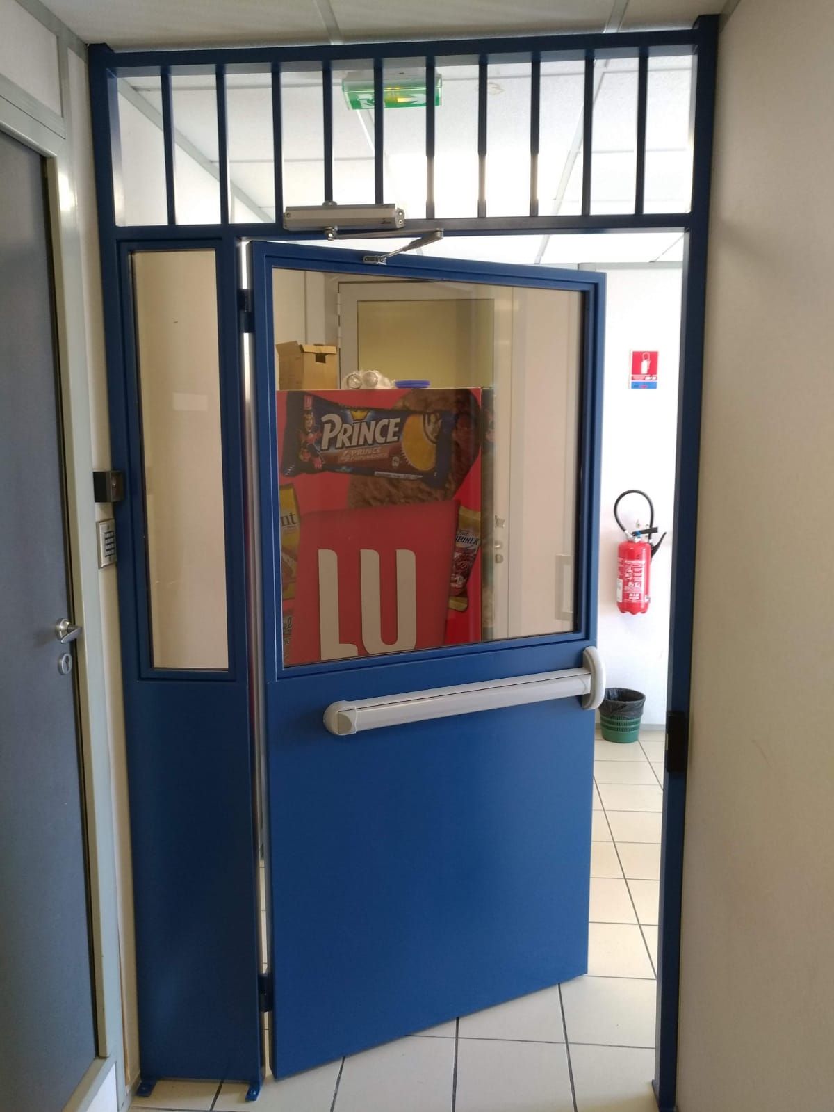 Une porte métallique, dotée d'une vitre et d'une barre antipanique horizontale.