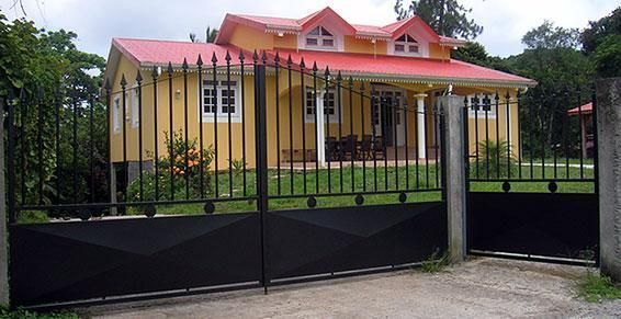 Une maison jaune au toit rouge derrière un portail et une clôture en métal noir.