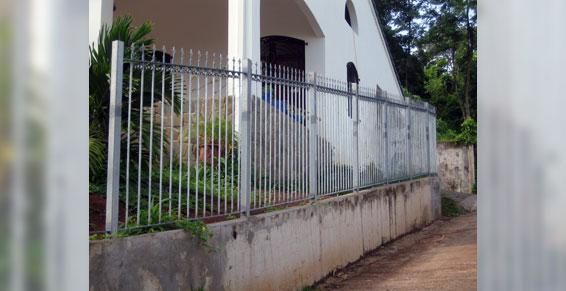 Un bâtiment blanc se dessine derrière une haute clôture métallique grise, fixée sur un muret de soutènement en béton.