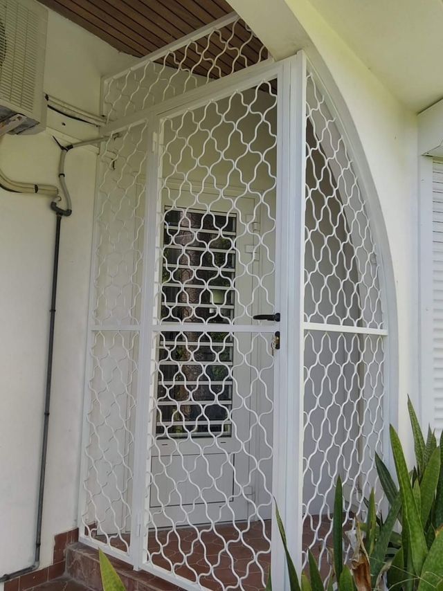 Une grille de protection en métal blanc orné d'un motif décoratif en maille ondulée, installé avant une entrée résidentielle.