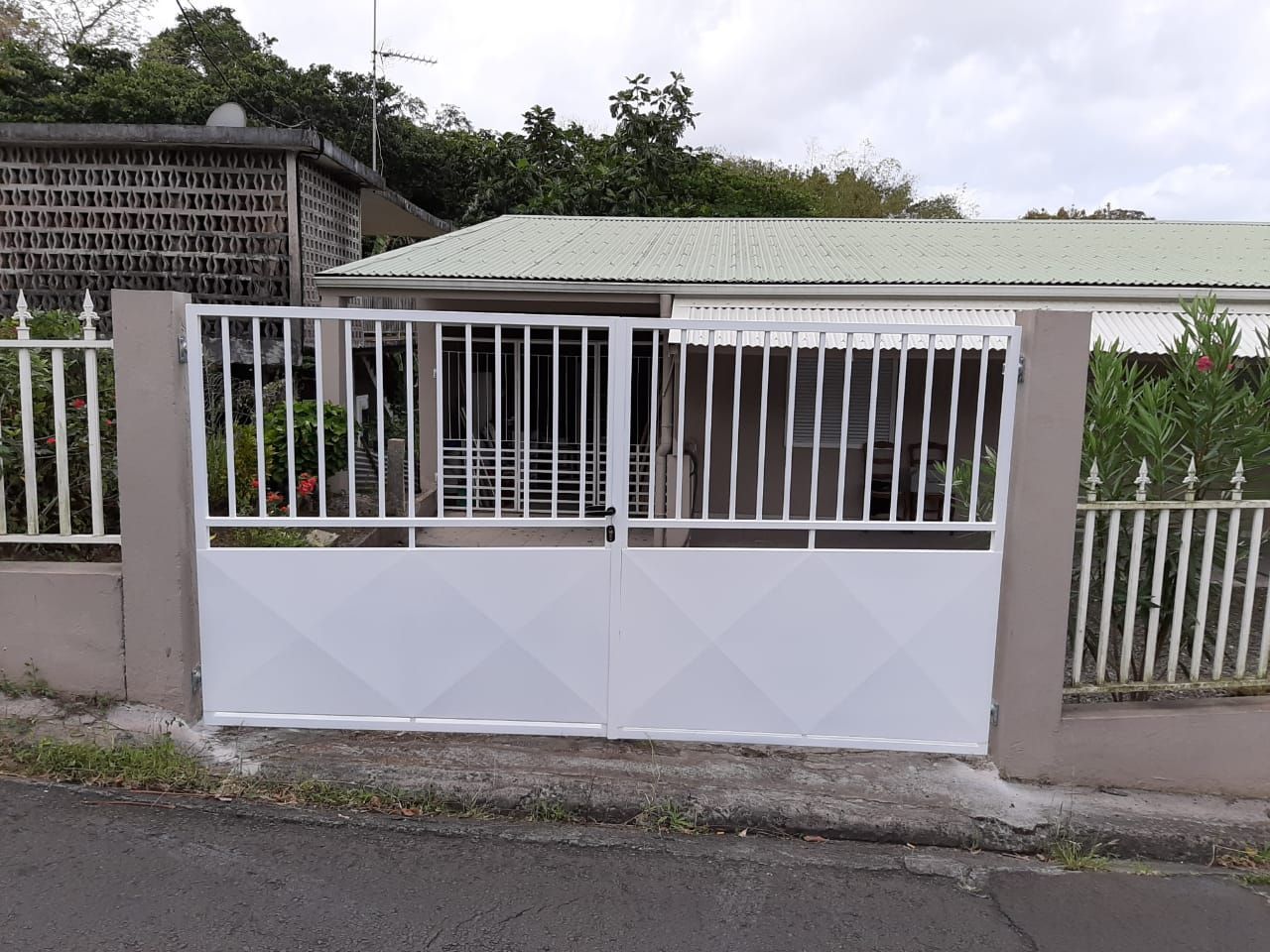 Un portail en métal blanc à panneaux à motifs losanges se dresse fermé devant une maison au toit vert et aux murs en béton.