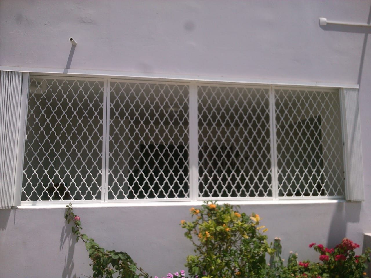 Une grille de protection blanche à mailles en losanges recouvre une grande fenêtre rectangulaire.