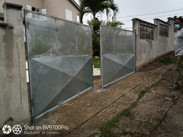 Un portail battant en métal gris, avec des bases à motifs en losange et des parties supérieures en treillis.