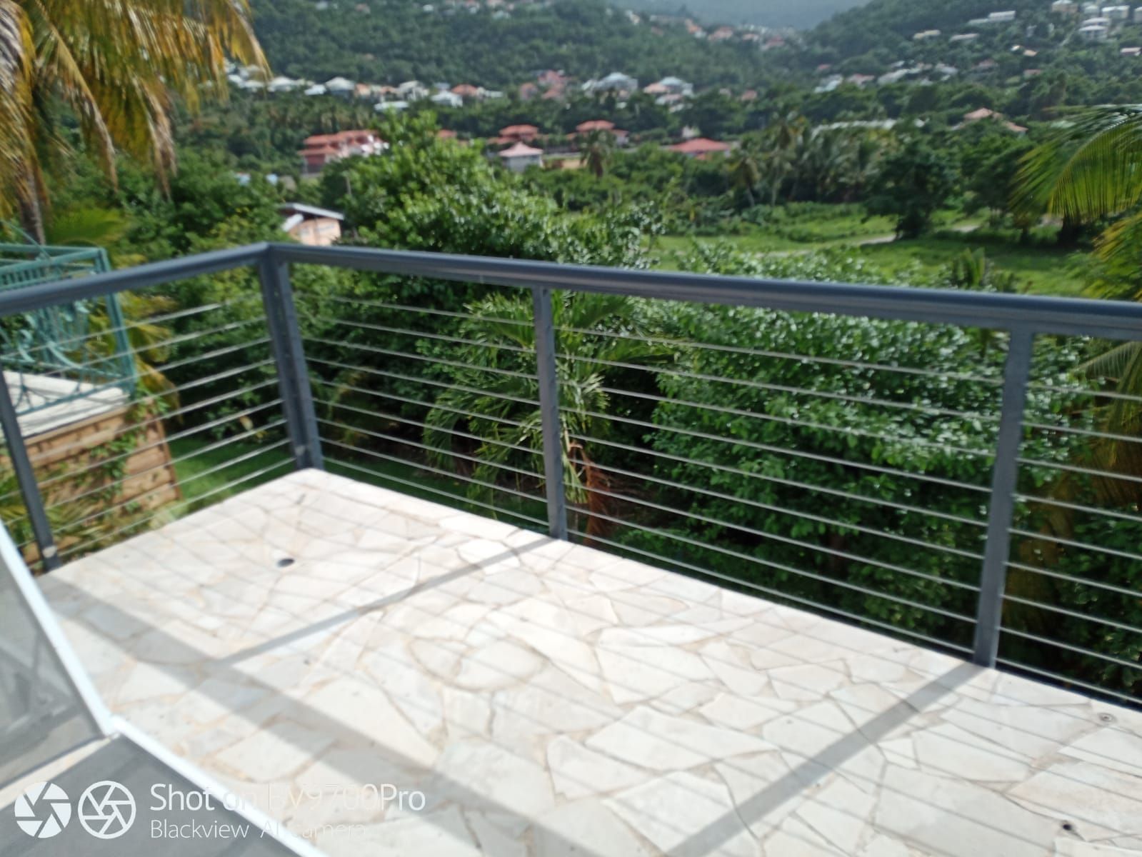 Un balcon avec un garde-corps en métal gris donne sur un paysage verdoyant luxuriant avec des maisons au loin.