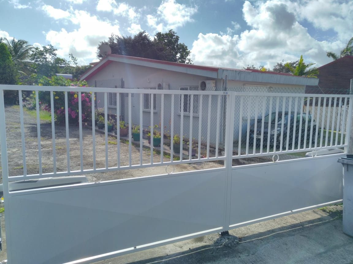 Un portail en métal blanc ouvre sur une cour où se dresse une maison.