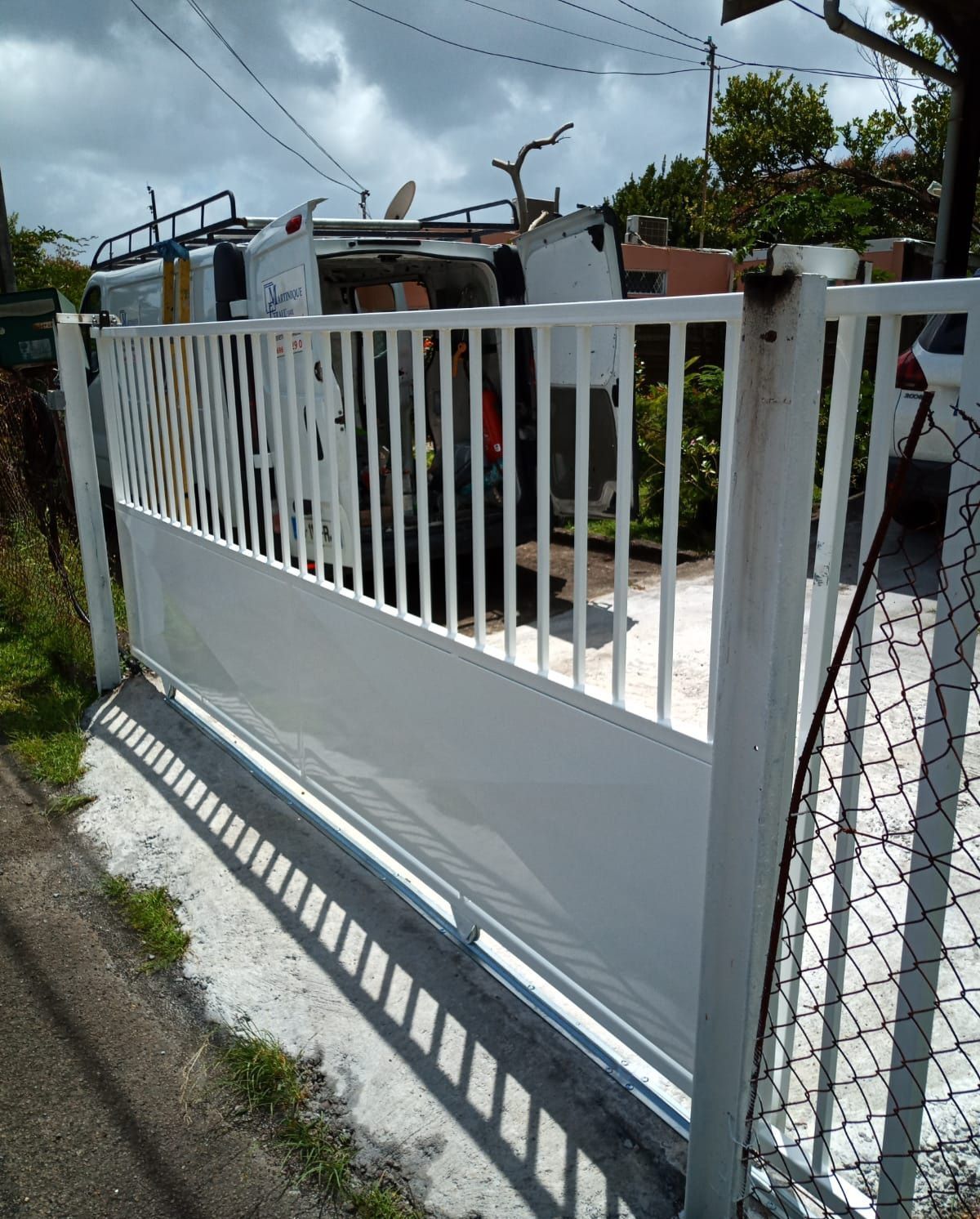 Un portail métallique blanc est fermé devant une camionnette blanche garée sur une allée en béton.