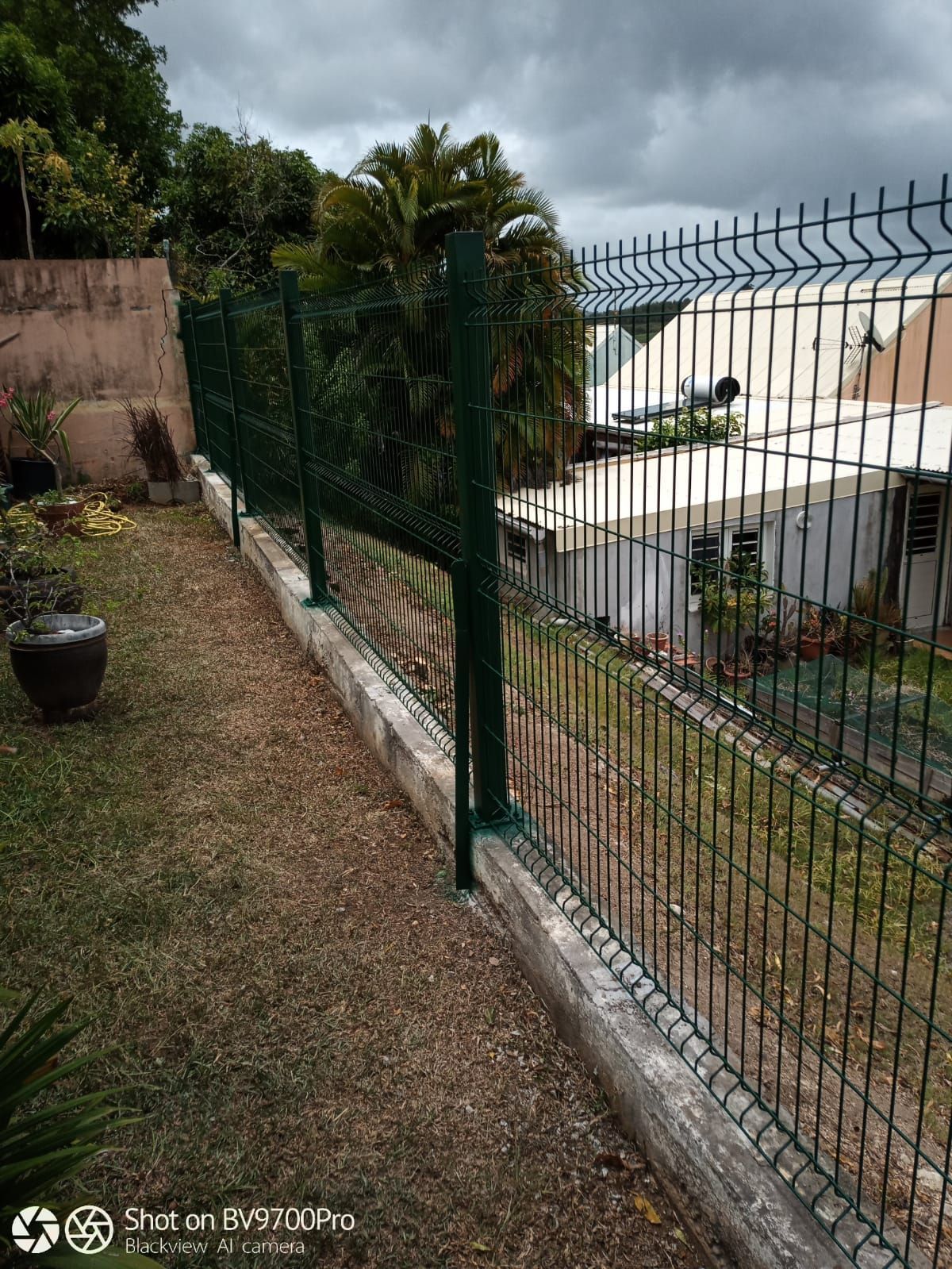Par temps nuageux, une clôture en fil de fer vert longe un mur de soutènement en béton qui borde une cour arrière gazonnée.