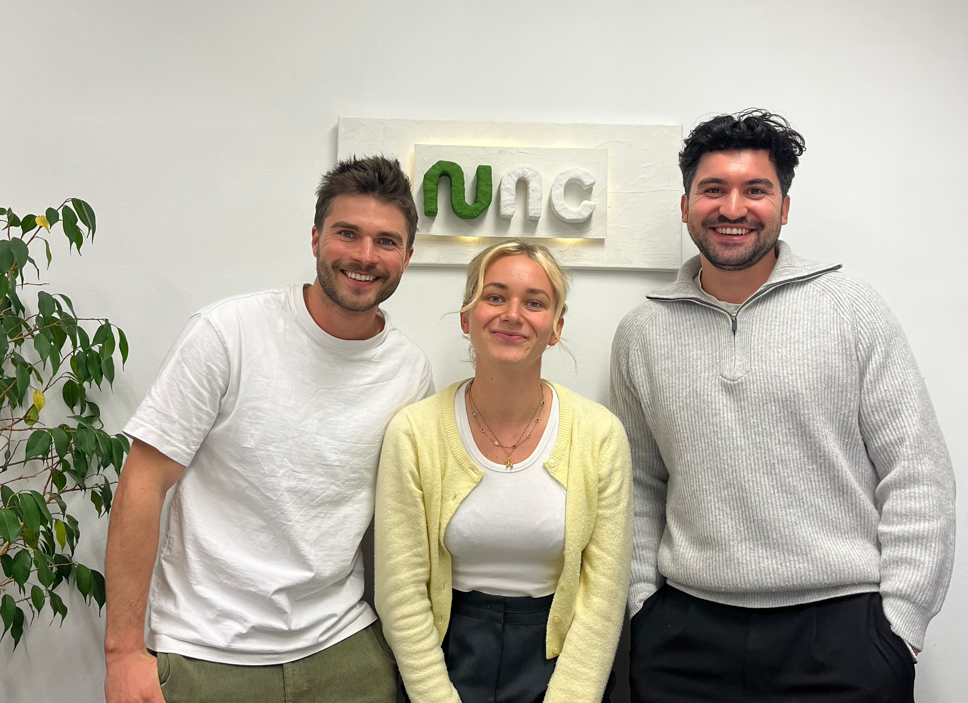 Zwei junge Männer und eine junge Frau in der Mitte. Sie stehen vor einer Wand und lachen in die Kamera