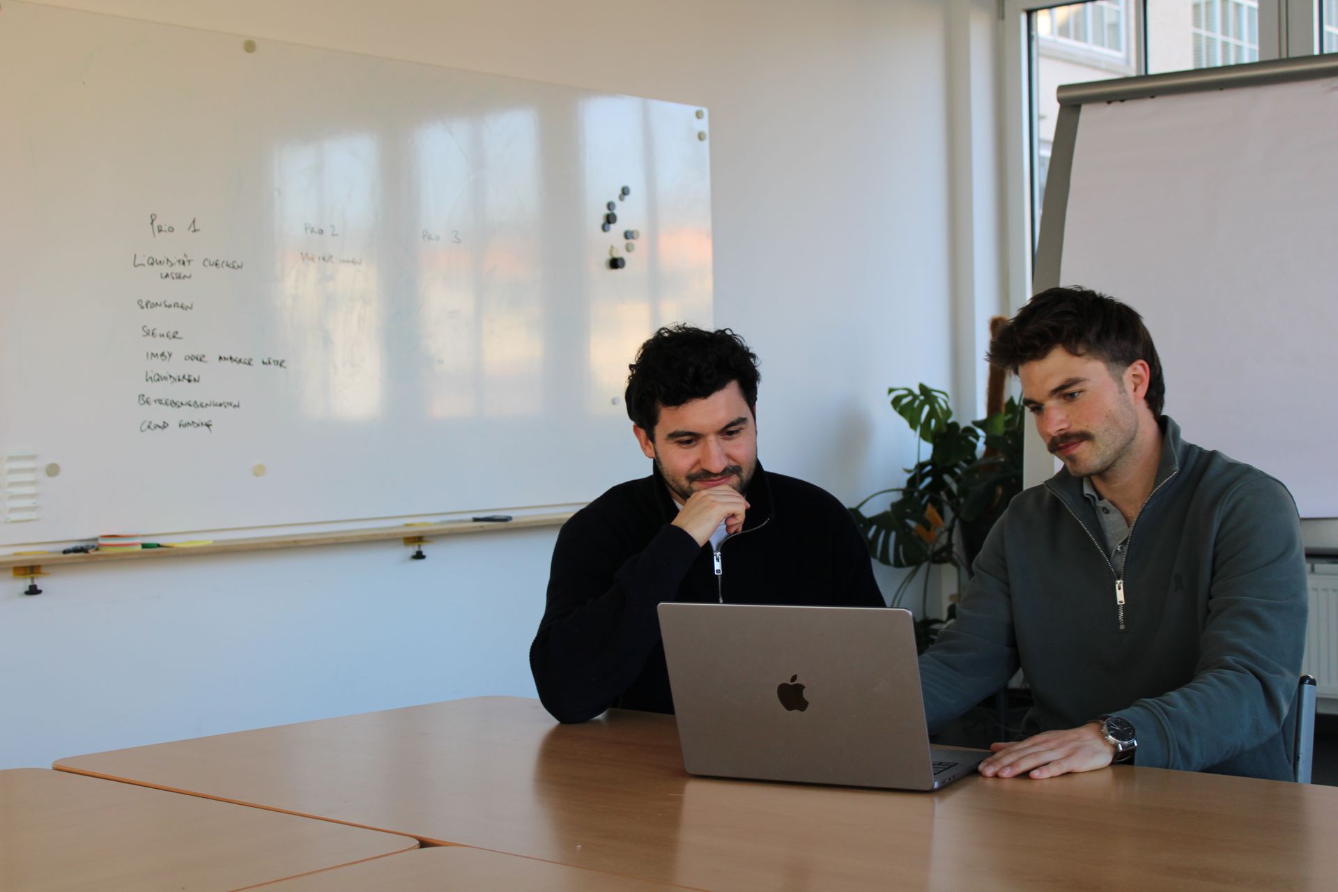 Zwei Männer sitzen an einem Tisch und schauen auf einen Laptop.