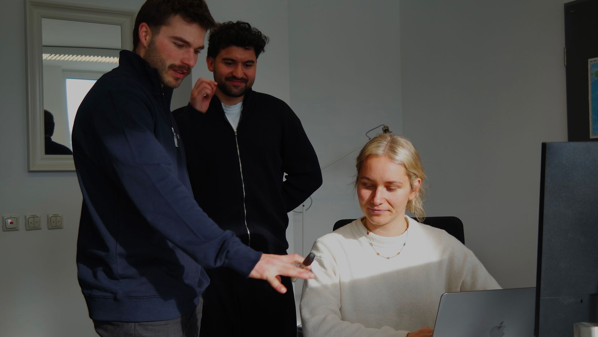 Zwei Kollegen stehen hinter einer sitzenden Person, die in einem Büro einen Laptop überprüft.