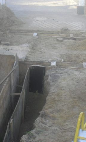 Baustelle mit Graben und hölzernen Stützwänden, die zu einem dunklen Tunnel führen.
