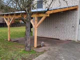 Ein hölzerner Carport ist an ein Backsteingebäude angebaut. Der Carport hat ein Metalldach und wird von Holzbalken getragen. Davor steht ein Baumstamm.