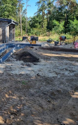 Eine Baustelle mit Gebäudefundament, Erde und Baumaschinen, eingebettet in ein Waldgebiet an einem sonnigen Tag.