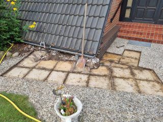 Die Dachfläche ist mit entfernten Steinplatten belegt, eine Schaufel lehnt am Gebäude. Garten, Kies und Gartenschlauch sind sichtbar.