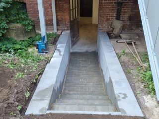 Eine Betontreppe führt hinunter zu einem Eingang, der von niedrigen Stützmauern flankiert wird. Im Hintergrund ist ein Backsteingebäude zu sehen.