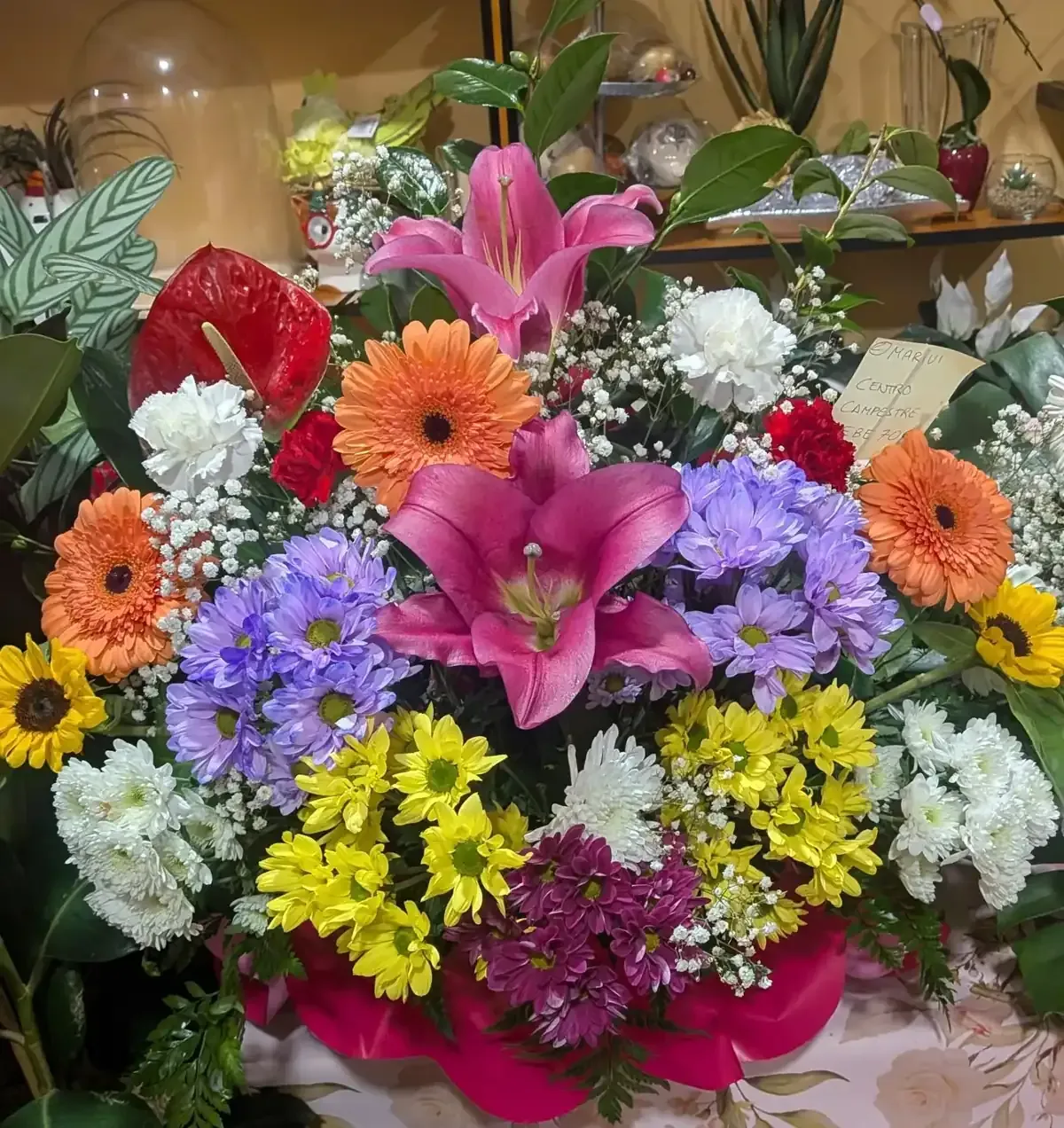 Arreglo floral mixto y vistoso con lirios rosas, margaritas naranjas y flores moradas y amarillas en un jarrón.