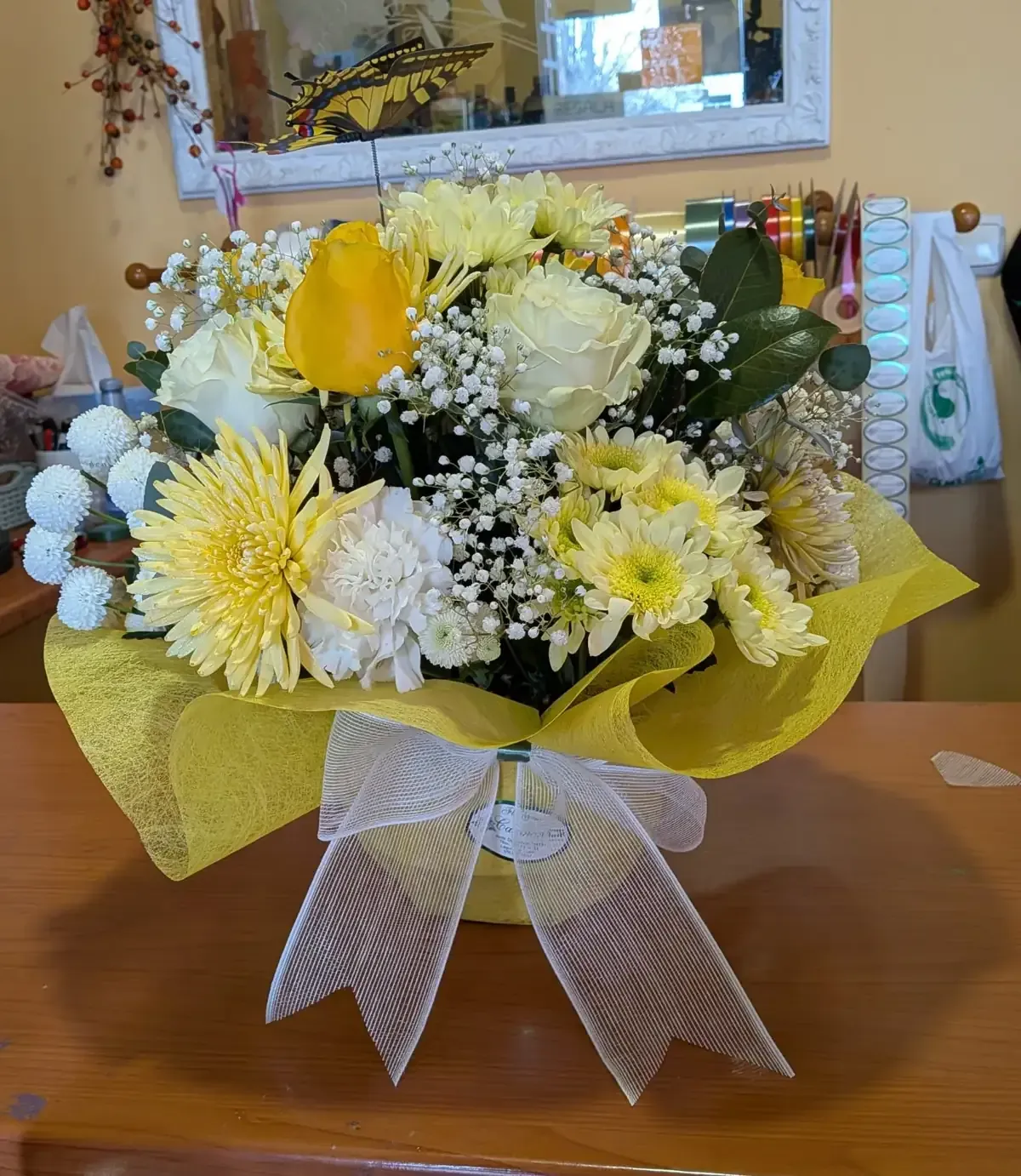 Ramo de flores amarillas y blancas con un lazo de cinta transparente sobre una mesa de madera.
