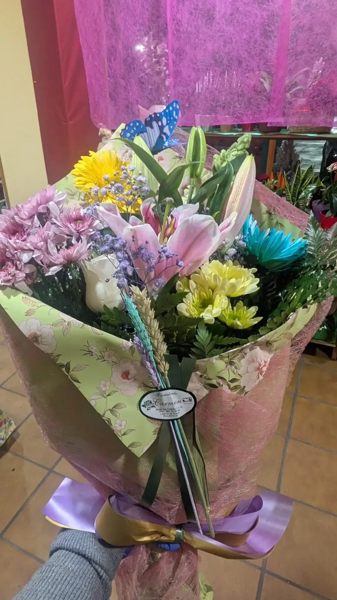 Un colorido ramo de flores variadas envuelto en papel rosa y atado con una cinta morada sobre un suelo de baldosas.