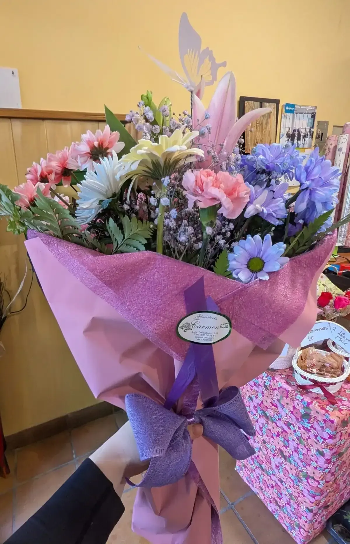 Ramo de flores rosas, moradas y blancas envuelto en papel rosa con un lazo morado, para llevar en la mano.