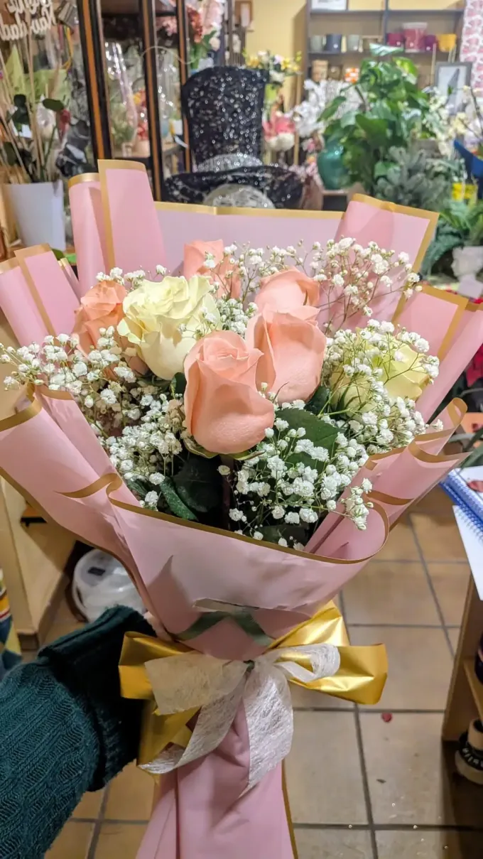 Ramo rosa y crema con rosas y paniculata, envuelto en papel y cinta de tonos pastel en una floristería.