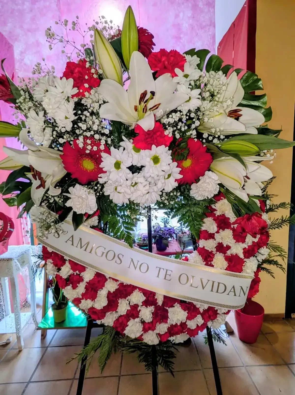 Gran corona fúnebre blanca y roja con lirios y una cinta que dice “AMIGOS NO TE OLVIDAN”.