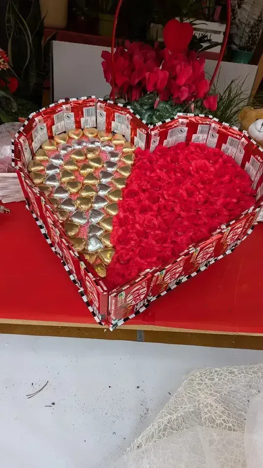 Caja de regalo en forma de corazón llena de flores rojas y bombones envueltos sobre una mesa roja.