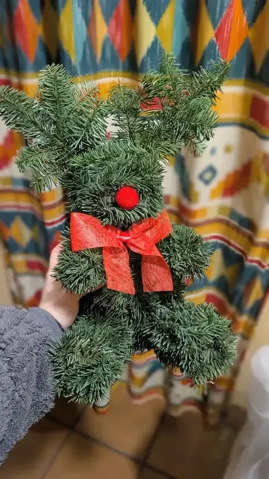 Oso de peluche de pino verde hecho a mano con un lazo rojo y una flor, sostenido frente a una cortina estampada.