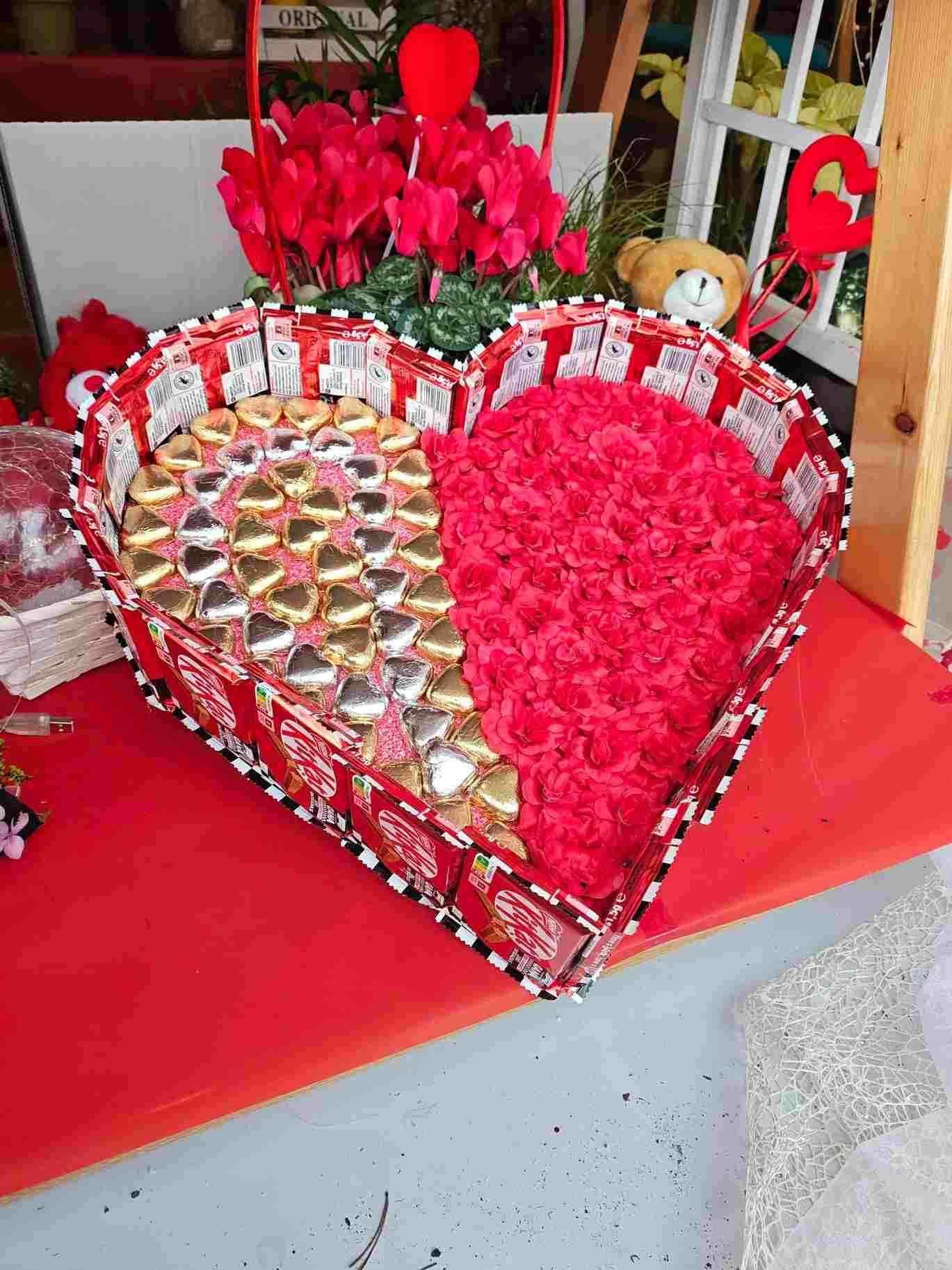 Arreglo floral rojo en forma de corazón y chocolates sobre una mesa, con rosas rojas detrás.