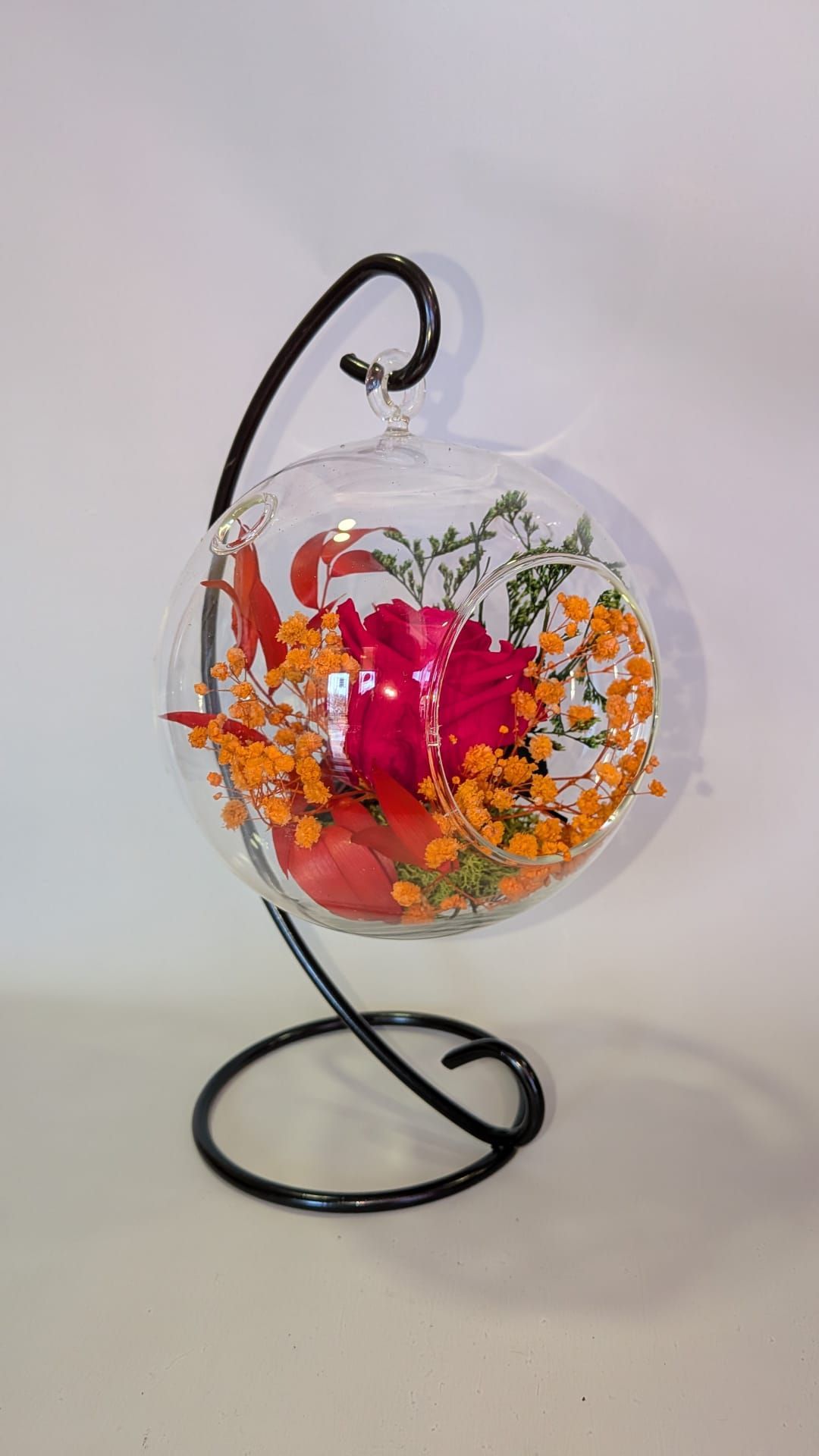 Adorno colgante de globo transparente con flores secas rojas y naranjas sobre una base de metal negro.