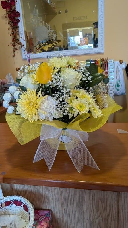 Un ramo de flores amarillas y blancas con cinta plateada sobre una mesa de madera cerca de un espejo.