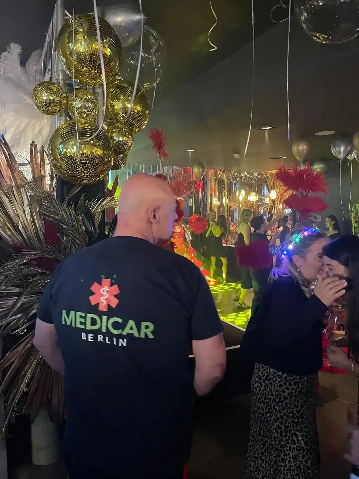 Ein Mann in einem T-Shirt von Medicar Berlin steht in einem überfüllten Raum.