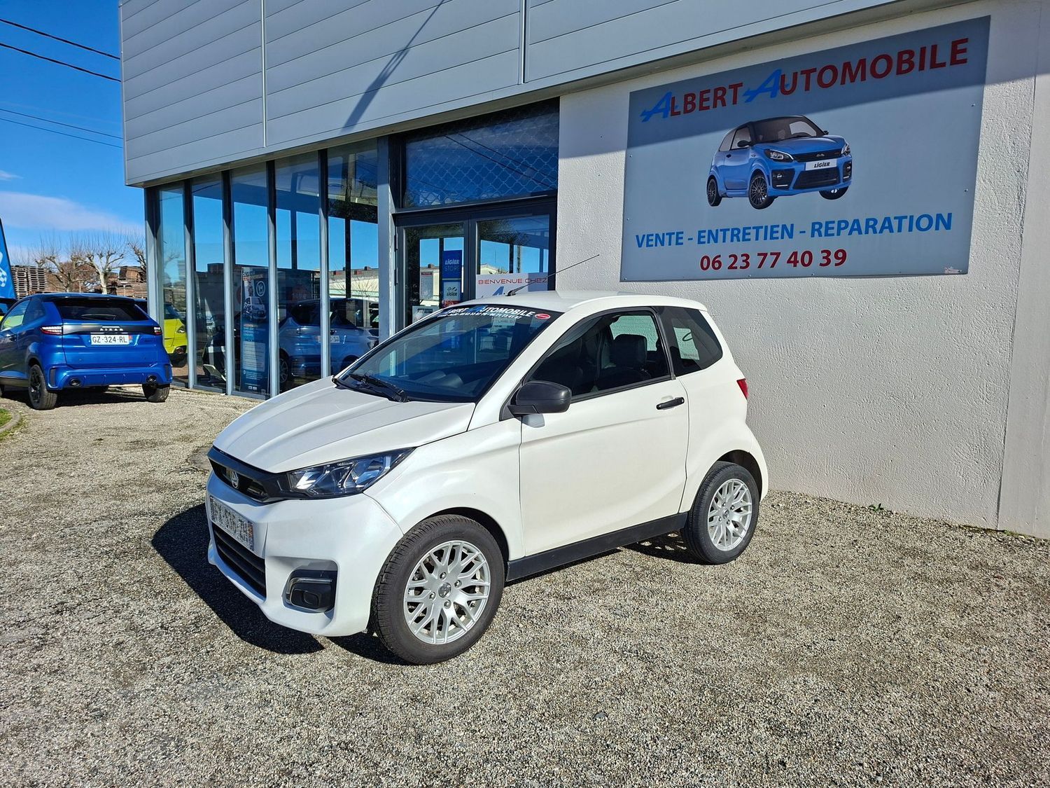 Une voiture compacte est garée devant un garage automobile avec une enseigne, une autre voiture se trouvant à proximité.
