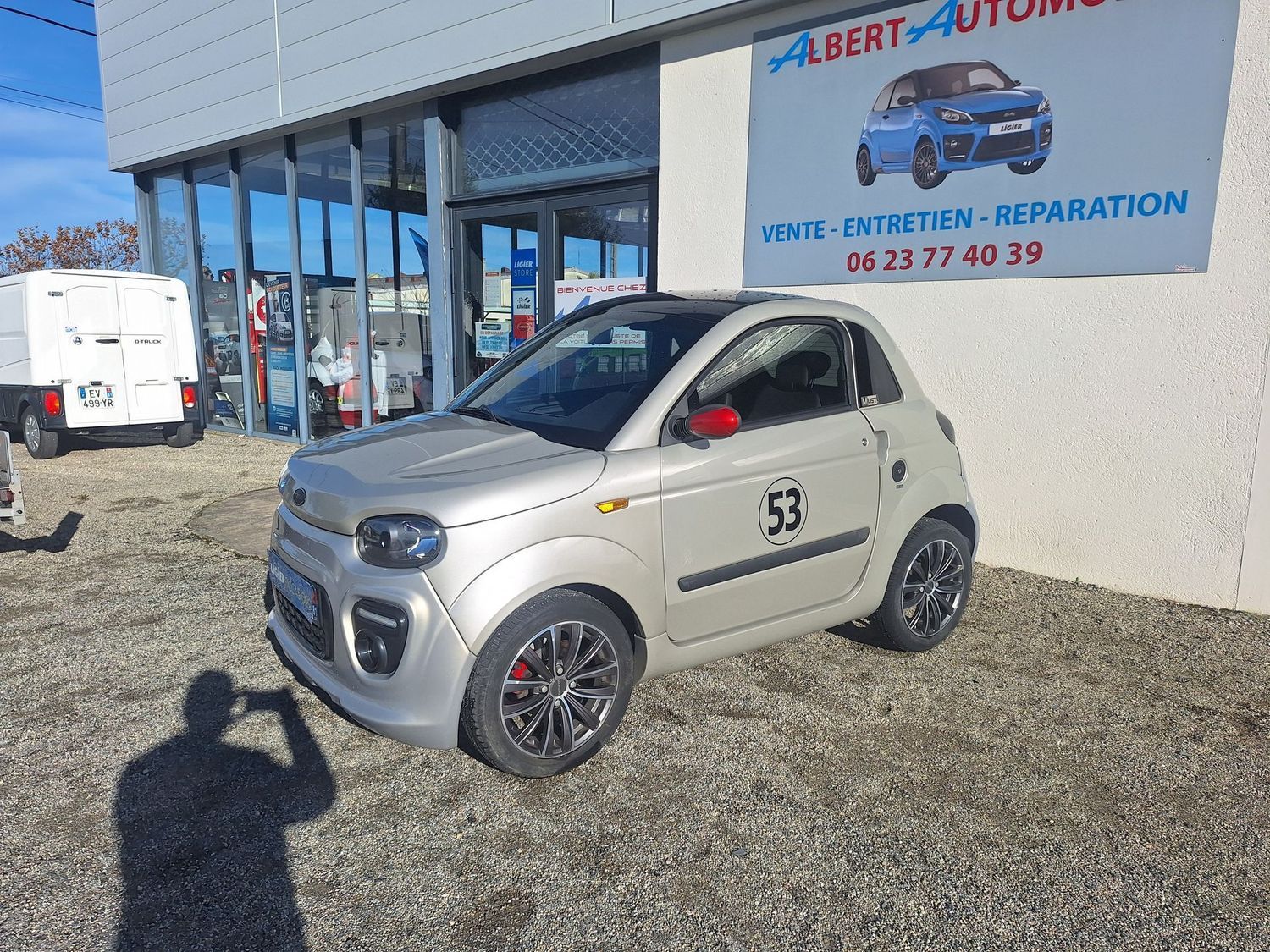Une petite voiture est garée devant un garage avec des panneaux de signalisation et une camionnette.
