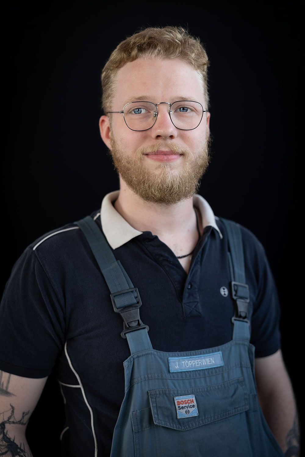 Ein Mann mit Bart und Brille trägt einen Overall und ein schwarzes Hemd.