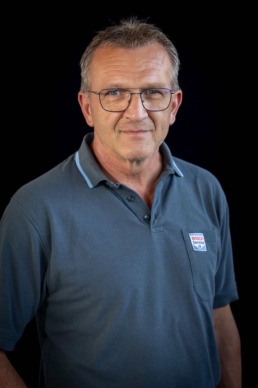 Ein Mann mit Brille und blauem Poloshirt steht vor einem schwarzen Hintergrund.