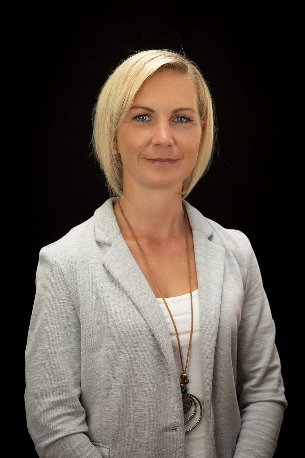 Eine Frau mit einer grauen Jacke und einer Halskette steht vor einem schwarzen Hintergrund.