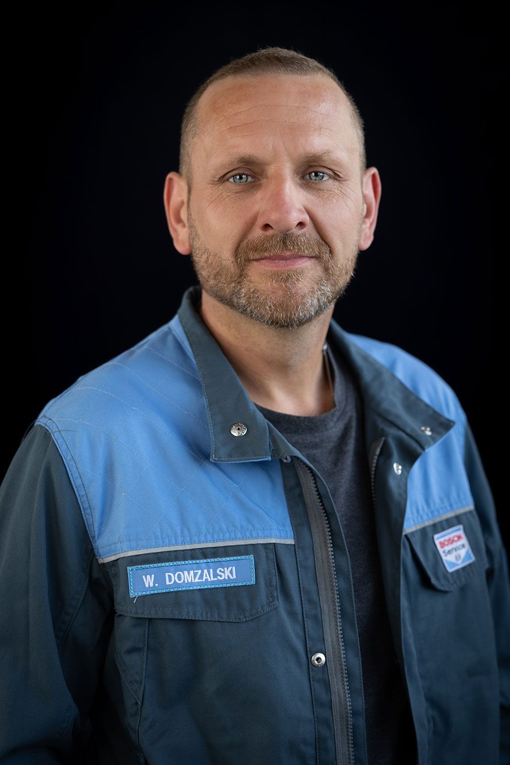 Ein Mann mit Bart trägt eine blaue Jacke und steht vor einem schwarzen Hintergrund.