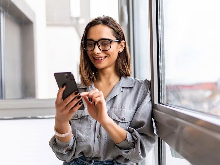 Eine Frau mit Brille steht neben einem Fenster und schaut auf ihr Handy.
