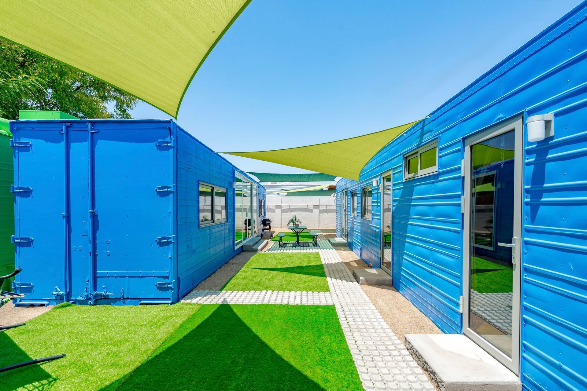 Blue shipping containers converted into buildings with a green lawn, a white walkway, and a yellow shade cover against a blue sky.