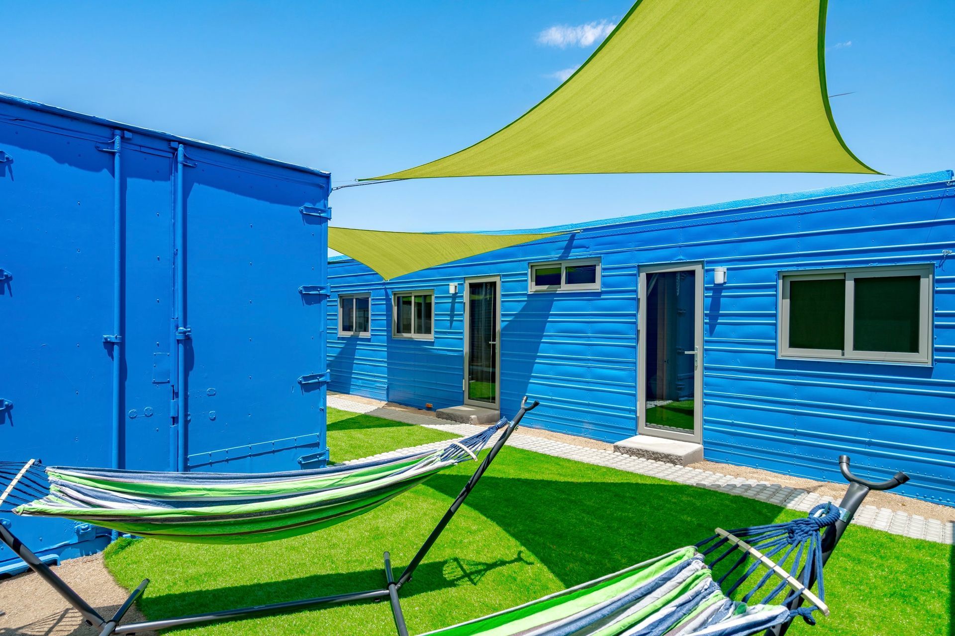 Blue structures with hammocks in a courtyard, shaded by green canopies. Sunny, outdoor setting.