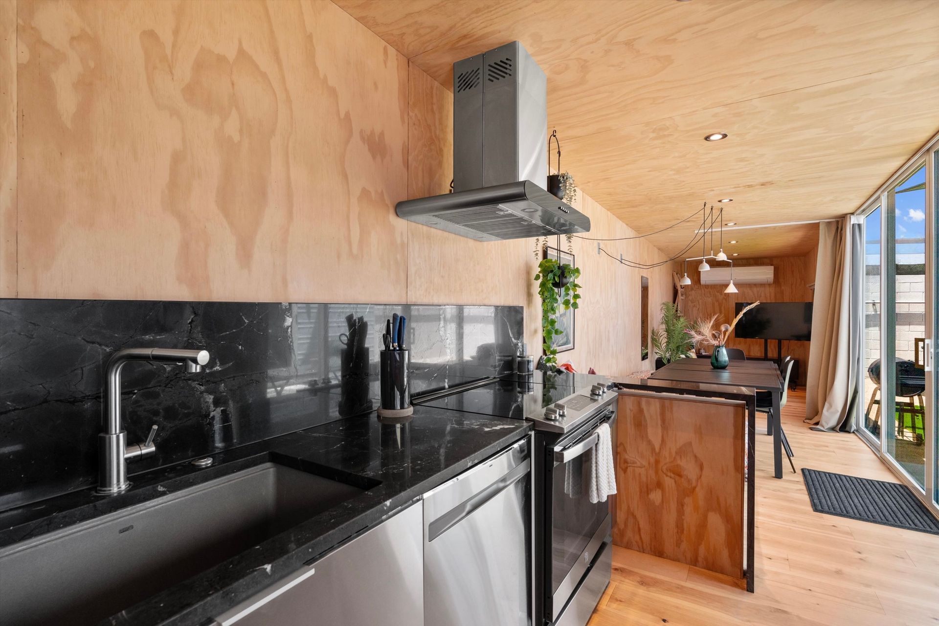 Modern kitchen with black countertop, stainless steel appliances, and wood paneling. Dining area visible, leading to a glass door.