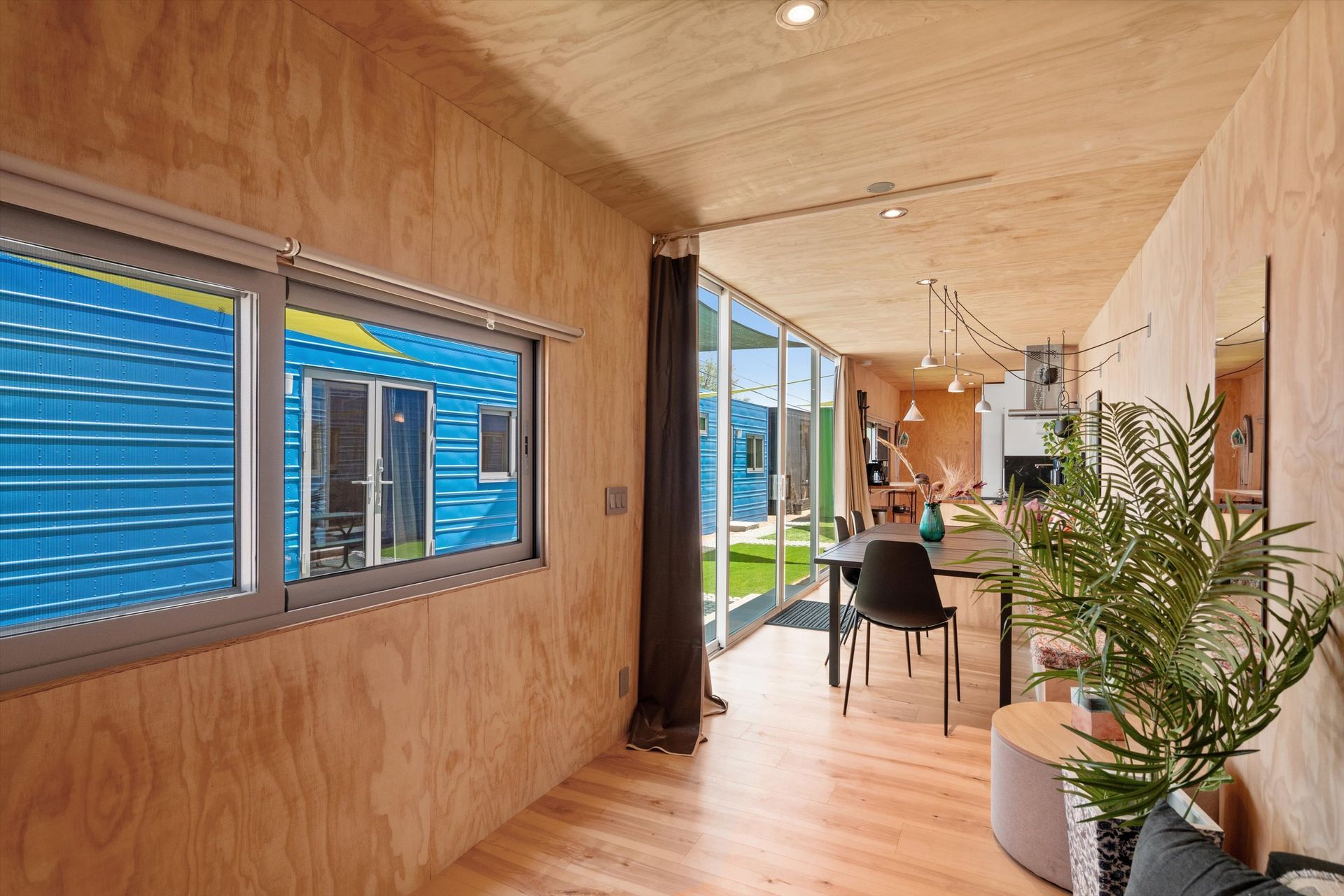 Interior view of a long, narrow room with wood paneling. Windows look out onto a blue building. A dining area with a table and chairs is in the distance.
