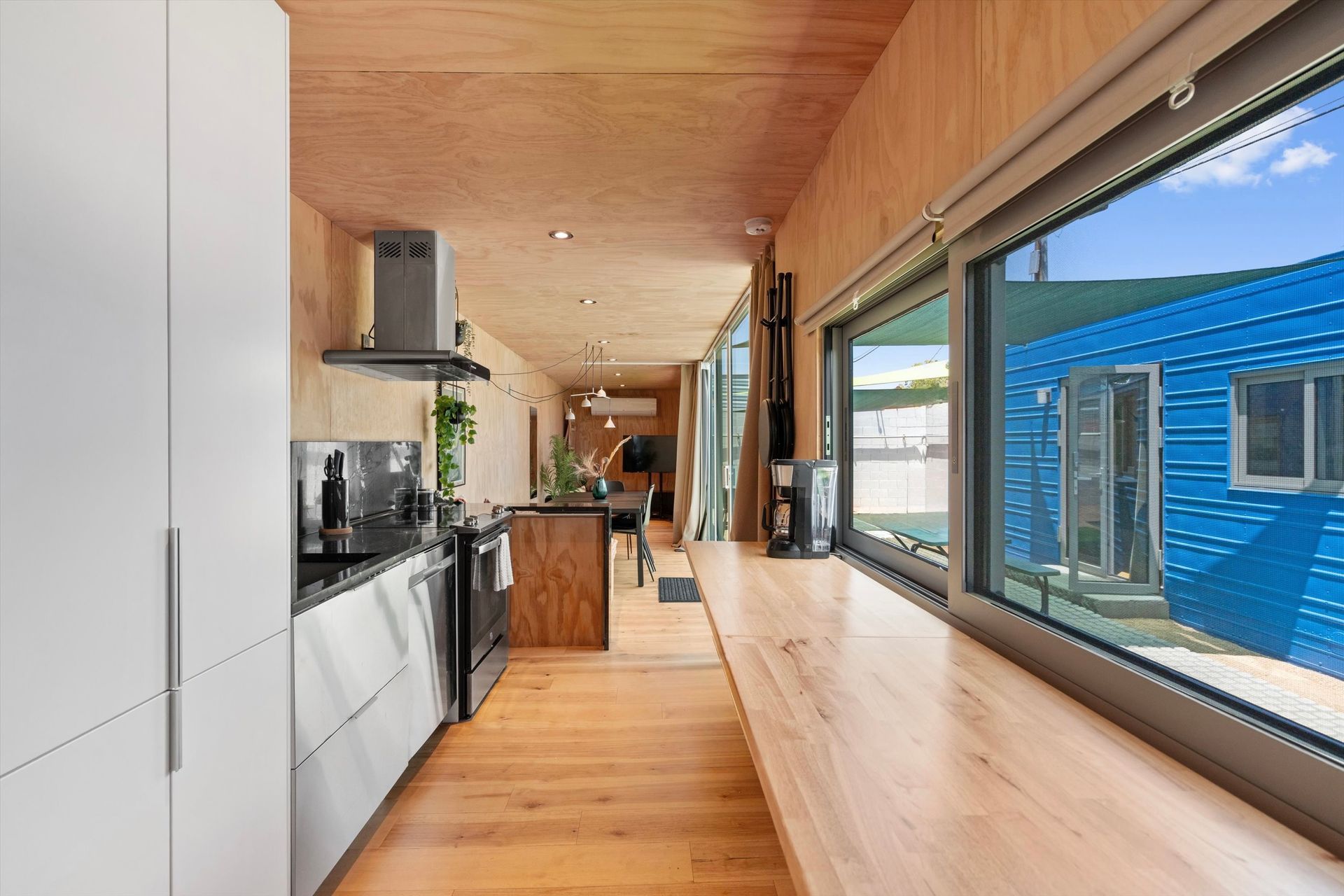 Modern kitchen with wood floors and walls, a large window overlooking a blue structure. White cabinets and appliances are visible.