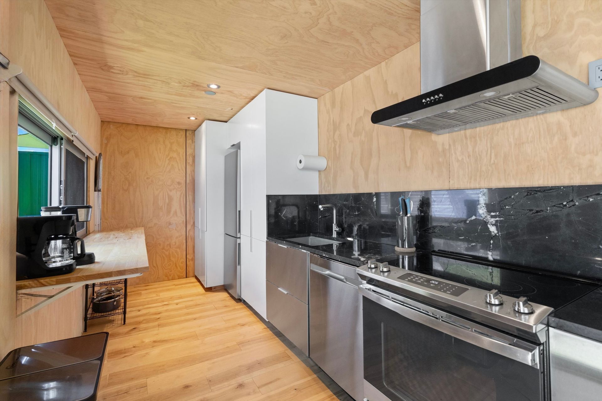 Modern kitchen with wood paneling, stainless steel appliances, and a black backsplash. A window lets in light.