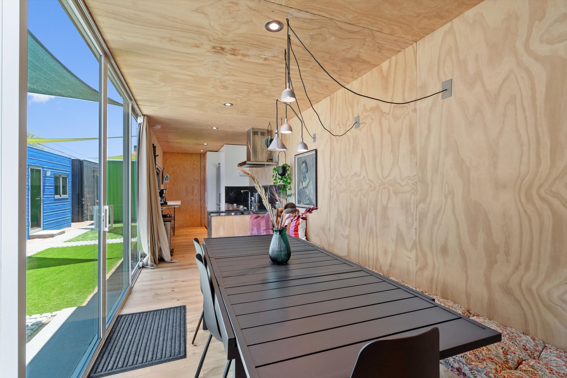 Interior view of a modern dining room with a long black table, wood panel walls, and a glass door leading to a yard.